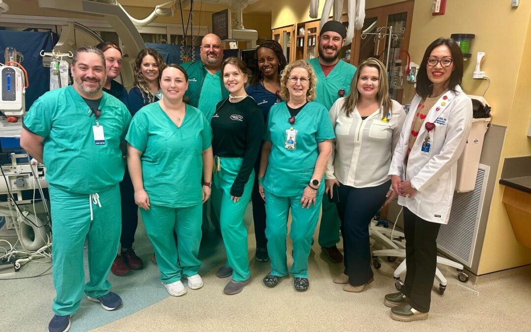 A group of healthcare professionals, some in green scrubs and others in business attire, pose and smile together in a hospital or medical facility Medical equipment is visible in the background