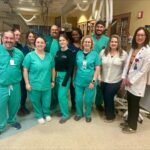 A group of healthcare professionals, some in green scrubs and others in business attire, pose and smile together in a hospital or medical facility Medical equipment is visible in the background