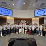 A group of people stands together in a council chamber, posing for a photo Many are holding certificates, and all wear name badges Two large screens display the logo for Palm Coast in the background