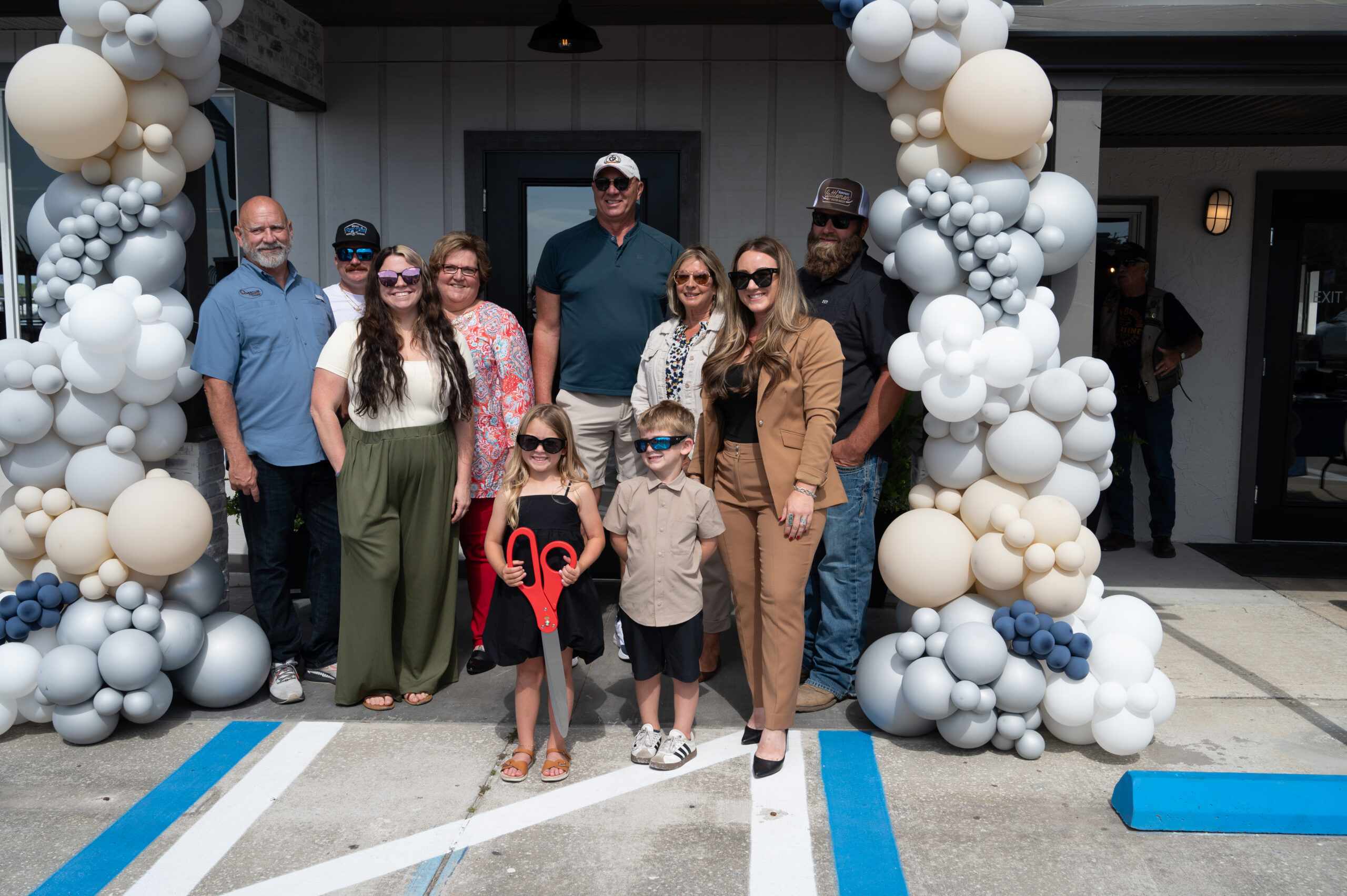 A group of adults and two children pose for a photo outside a building, surrounded by balloon decorations The children stand in front, one holding large ceremonial scissors Everyone is smiling on a sunny day