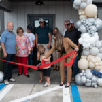 A group of people stand outside a building decorated with white and gray balloons, as a young boy and a woman in a suit cut a red ribbon during a ribbon cutting ceremony Others watch and smile