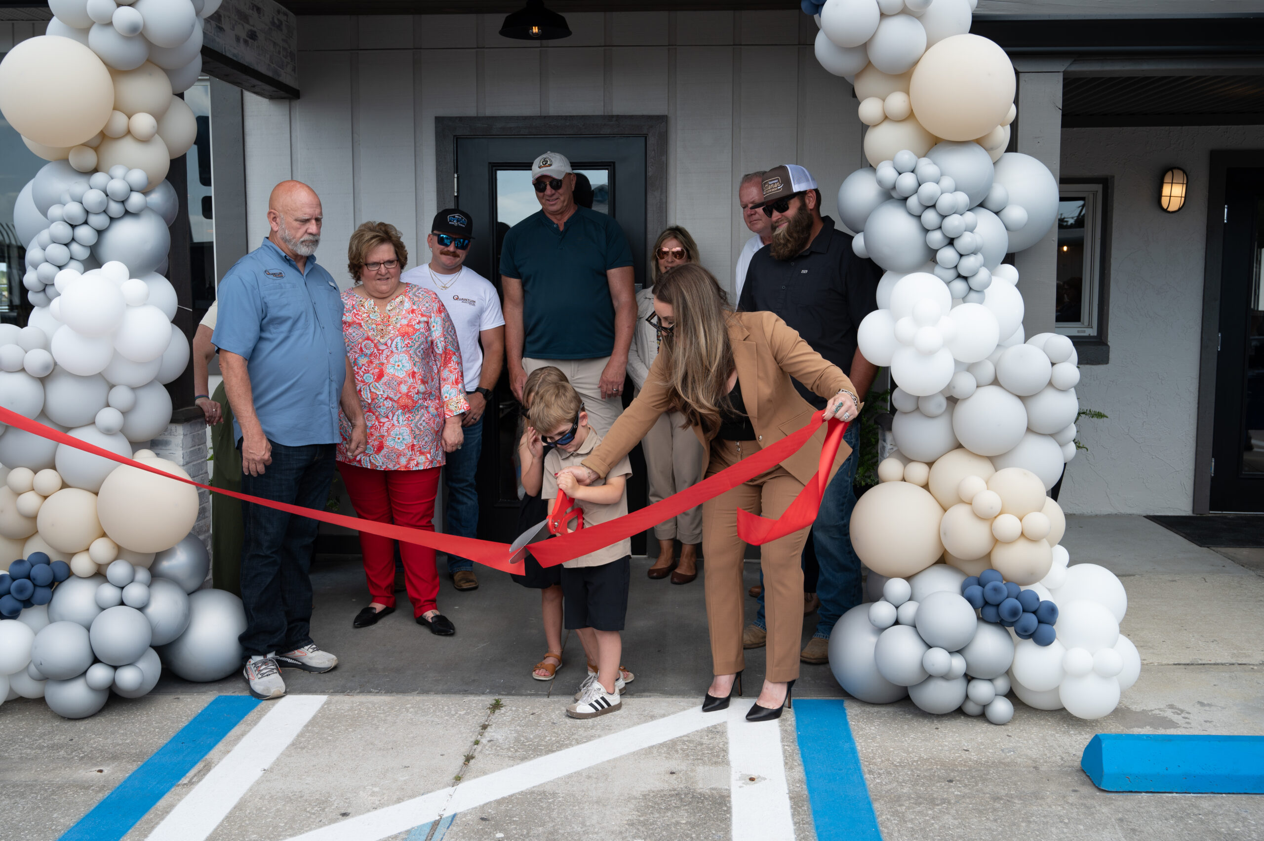 A group of people stand outside a building decorated with white and gray balloons, as a young boy and a woman in a suit cut a red ribbon during a ribbon cutting ceremony Others watch and smile