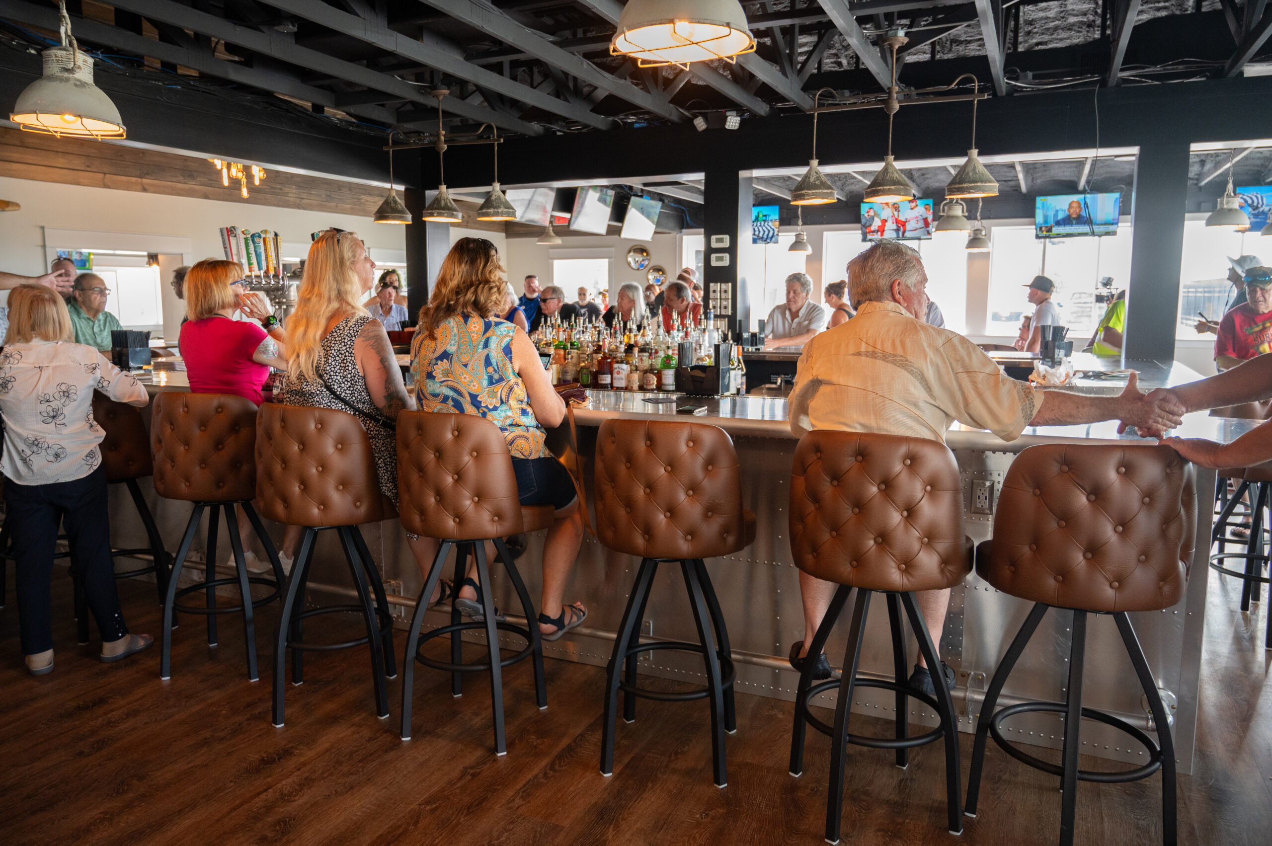 People sit on cushioned barstools around a large, modern bar, chatting and enjoying drinks Several TV screens hang above the bar, and more people stand and mingle in the background