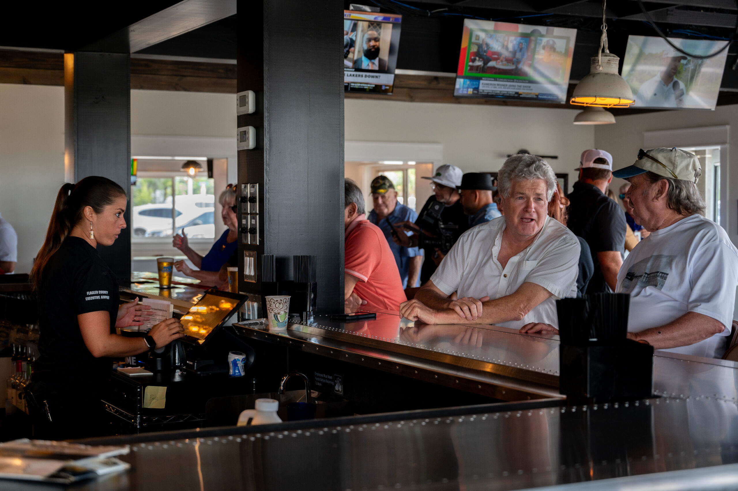 A bartender serves drinks to patrons seated at a busy bar Two men in white shirts converse at the counter, while several people watch sports on TVs mounted above The atmosphere is lively and casual