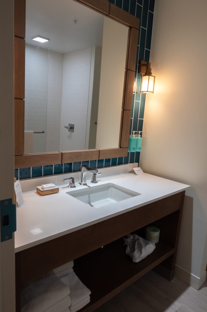Modern bathroom vanity with a rectangular sink, wooden framed mirror, wall sconce, blue tile backsplash, toiletries on the counter, and towels neatly stacked on the lower shelf Shower visible in the background