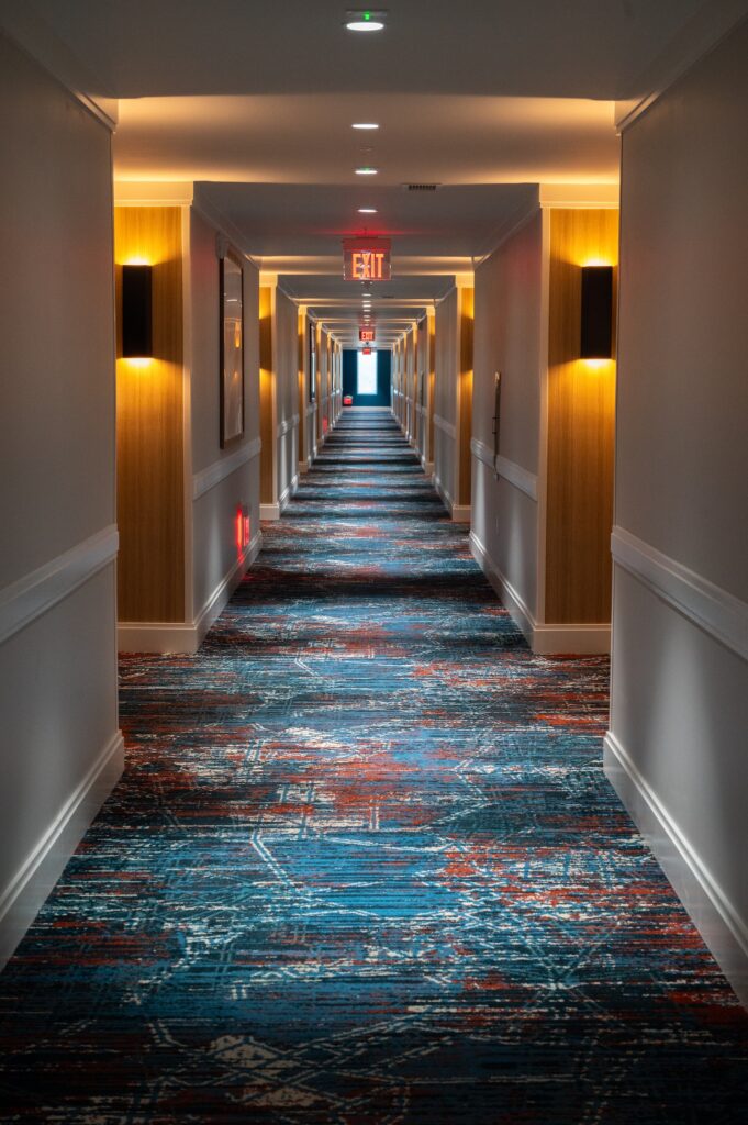 A long, brightly lit hotel hallway with colorful patterned carpet, wooden wall panels, and wall lights An exit sign is visible at the far end, with daylight shining through an open door