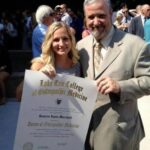 A young woman holding a Lake Erie College of Osteopathic Medicine diploma stands next to an older man in a suit, both smiling at an outdoor graduation event with people in the background