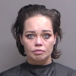 A woman with short, dark hair and striking light colored eyes looks directly at the camera with a neutral expression She is wearing a dark smock, and the background is plain gray