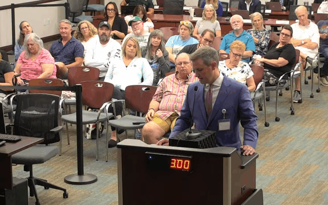 A man in a suit speaks at a podium displaying “:” in a meeting room filled with seated people who are watching and listening attentively The setting appears to be a public or community meeting