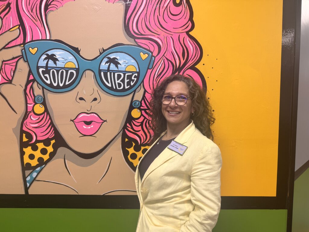 A woman in a light yellow blazer stands smiling in front of a colorful mural of a woman with pink hair and large sunglasses that say 