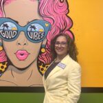 A woman in a light yellow blazer stands smiling in front of a colorful mural of a woman with pink hair and large sunglasses that say "GOOD VIBES," with palm trees and oceans reflected in the glasses
