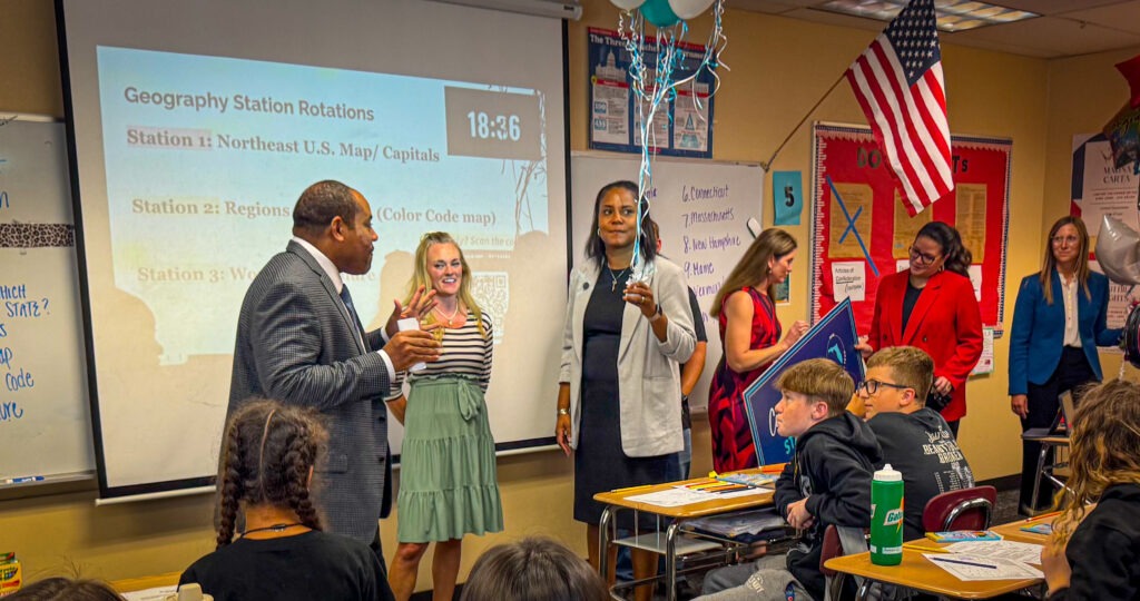 A group of teachers and students interact in a classroom A timer and geography station instructions are projected on a screen, and balloons are held by a woman standing near the center The class appears engaged and attentive