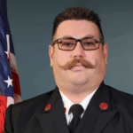 A man in a dark uniform with a large mustache and glasses stands in front of a U S flag, looking directly at the camera against a plain background