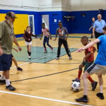 Children and adults play an energetic indoor soccer game in a gymnasium The players are focused on the ball, while others watch and cheer along the sidelines The atmosphere is lively and engaging