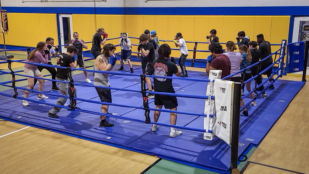 A group of people practice boxing techniques in pairs inside a large boxing ring, each wearing athletic gear and facing partners, with a coach observing in a gym setting