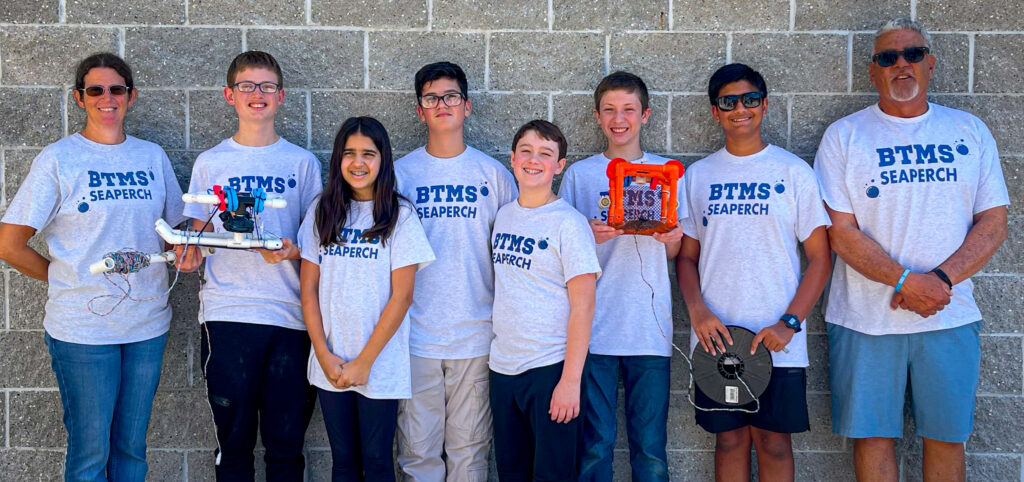 Seven students and two adults stand in front of a gray brick wall, all wearing matching "BTMS SEAPERCH" shirts Two students hold robotics equipment and everyone is smiling at the camera Seven students and two adults stand in front of a gray brick wall, all wearing matching "BTMS SEAPERCH" shirts Two students hold robotics equipment and everyone is smiling at the camera