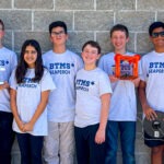 Seven students and two adults stand in front of a gray brick wall, all wearing matching "BTMS SEAPERCH" shirts Two students hold robotics equipment and everyone is smiling at the camera