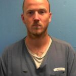 A man with a shaved head and short beard wearing a gray prison uniform stands against a plain blue wall, looking directly at the camera with a neutral expression