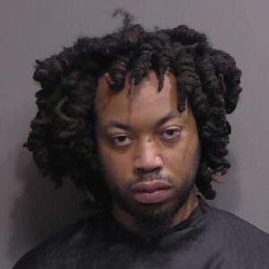 A man with short, twisted dreadlocks and facial hair looks directly at the camera with a neutral expression, wearing a dark colored garment against a plain background