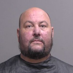 A middle aged, bald man with a beard is facing the camera for a mugshot, wearing a dark drape around his shoulders against a plain, neutral background