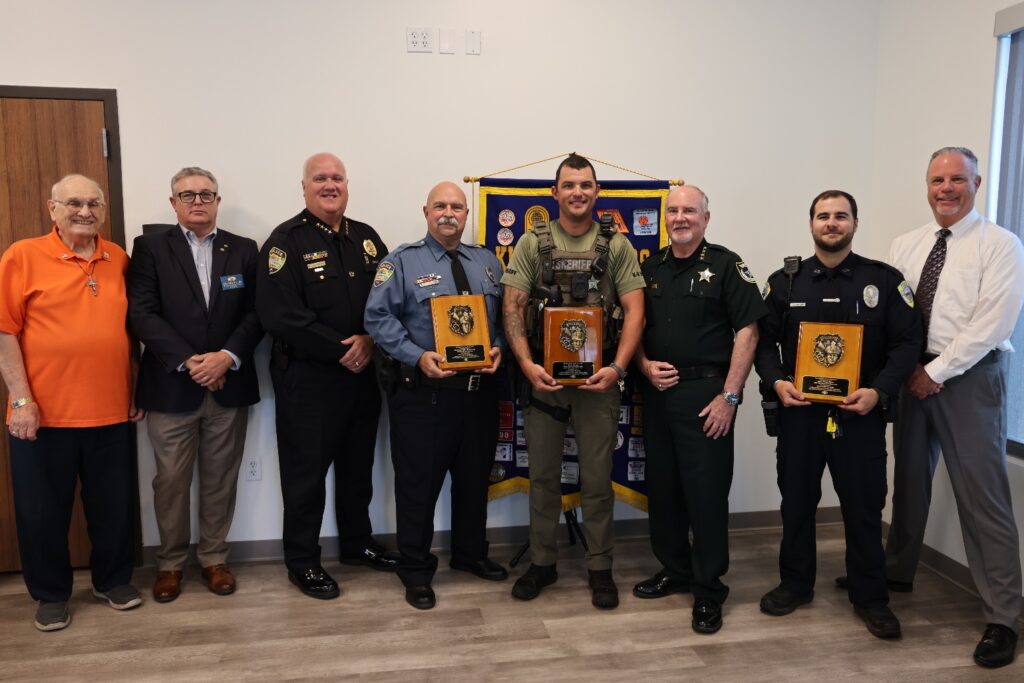 Eight men stand indoors, four in police or sheriff uniforms holding plaques, and four in business or casual attire They pose in front of a banner and smile at the camera, suggesting an award or recognition event