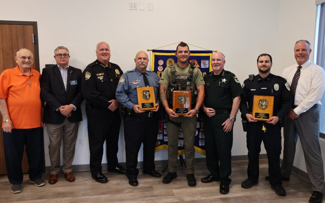 Eight men stand indoors, four in police or sheriff uniforms holding plaques, and four in business or casual attire They pose in front of a banner and smile at the camera, suggesting an award or recognition event