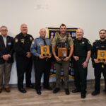 Eight men stand indoors, four in police or sheriff uniforms holding plaques, and four in business or casual attire They pose in front of a banner and smile at the camera, suggesting an award or recognition event
