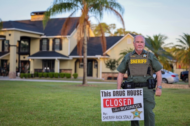 A sheriff stands in front of a house and palm trees, holding a sign that reads, "This DRUG HOUSE CLOSED FOR BUSINESS by Sheriff Rick Staly