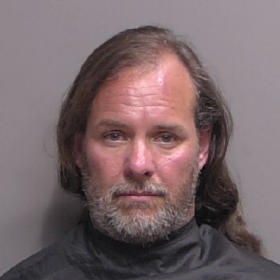 A middle aged man with long brown hair and a gray beard is facing the camera against a plain background, wearing a black covering around his shoulders