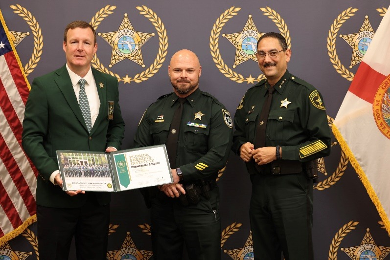 Three uniformed law enforcement officers stand in front of a backdrop with gold star emblems One holds a certificate, another poses in the center, and a third stands to the right The Florida state flag is partially visible