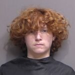 A young person with curly red hair and pale skin faces the camera with a neutral expression, wearing a dark garment against a plain gray background, evoking the subtle calm of Palm Coast