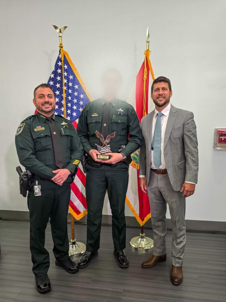 Three men stand indoors; one in a suit and two in sheriff uniforms The center person's face is blurred, holding an eagle trophy Behind them are the U S and Florida state flags against a plain wall