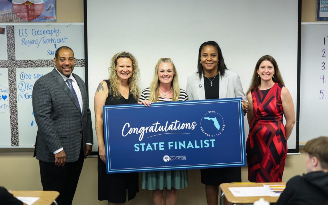 Five adults stand in a classroom holding a large blue sign that reads “Congratulations State Finalist” with a Florida outline, while students sit at desks in the foreground