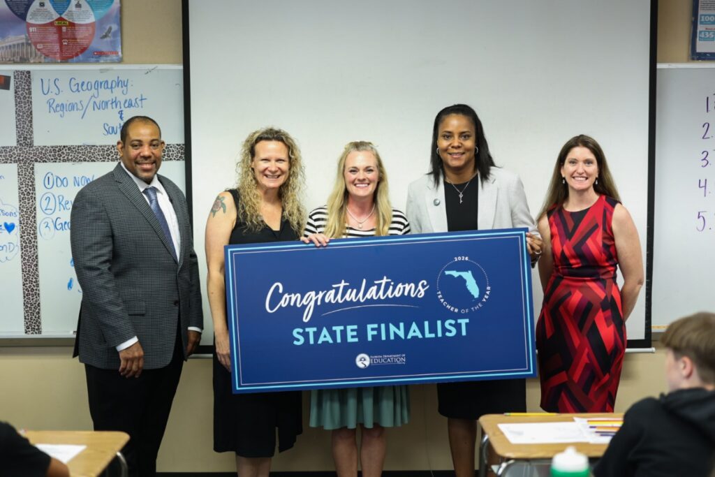 Five adults stand in a classroom, smiling and holding a large blue sign that reads "Congratulations State Finalist" with the outline of Florida and the Department of Education logo