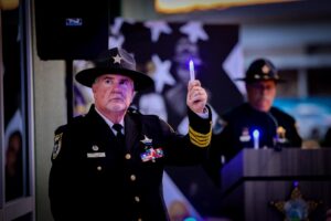 A uniformed police officer holds up a glowing blue light while standing indoors Another officer stands blurred at a podium in the background with a backdrop of stars behind them