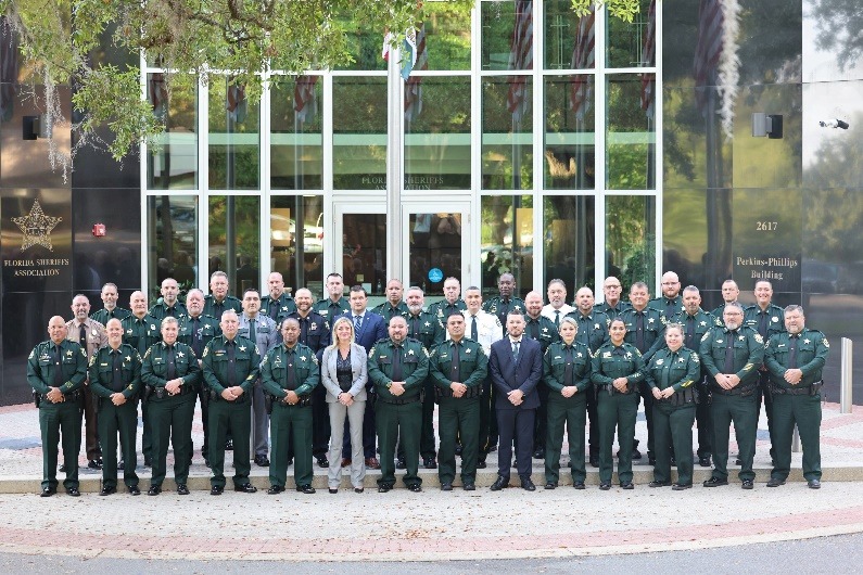 A large group of uniformed law enforcement officers and a few individuals in formal attire stand in rows outside a building with glass doors and the Florida Sheriffs Association sign