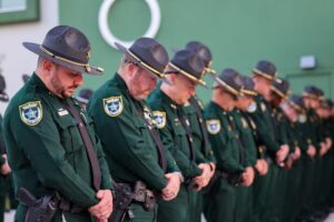 A group of uniformed law enforcement officers stand in a row with heads bowed and hands clasped, appearing to observe a moment of silence outside a green building
