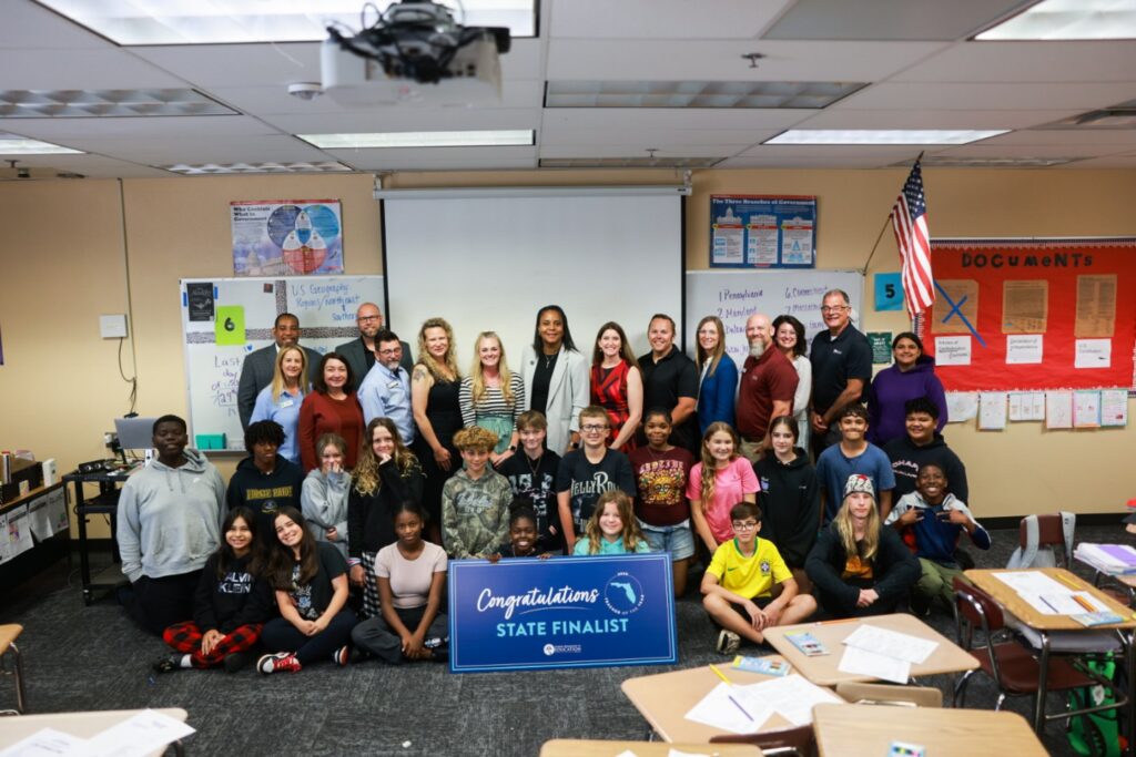 A diverse group of students and adults pose together in a classroom, some standing and some sitting, holding a banner that reads “Congratulations State Finalist ” Classroom posters and an American flag are visible in the background