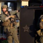 Two armed law enforcement officers in tactical gear stand beside an armored vehicle, one pointing while the other faces away The word “SHERIFF” is visible on a vest A house with large windows is in the background
