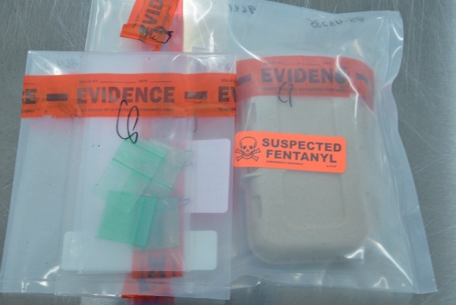 Two sealed evidence bags labeled "EVIDENCE"; one contains small green packets, the other contains a box with an orange "SUSPECTED FENTANYL" sticker and a skull symbol Both are on a metallic surface