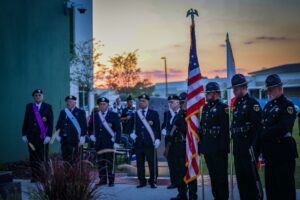 A group of uniformed veterans stands in formation during a ceremony at sunset Some hold flags, while others stand at attention A modern building and trees are visible in the background