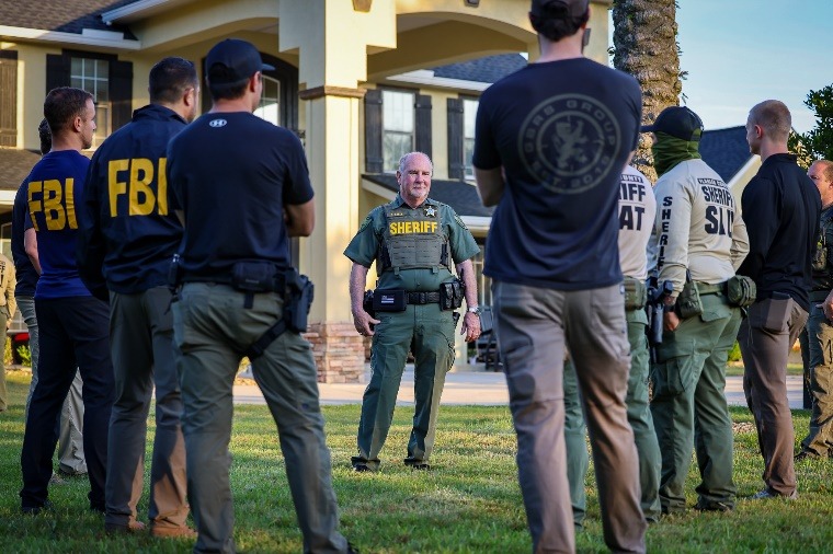 A sheriff stands in front of a group of law enforcement officers, some wearing shirts labeled FBI and SWAT, as they gather outside a suburban house on a lawn