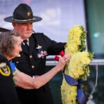 A police officer helps an elderly woman place a wreath of yellow flowers with blue ribbons during a memorial ceremony, while another officer stands nearby