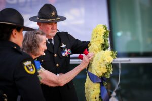 A police officer helps an elderly woman place a wreath of yellow flowers with blue ribbons during a memorial ceremony, while another officer stands nearby
