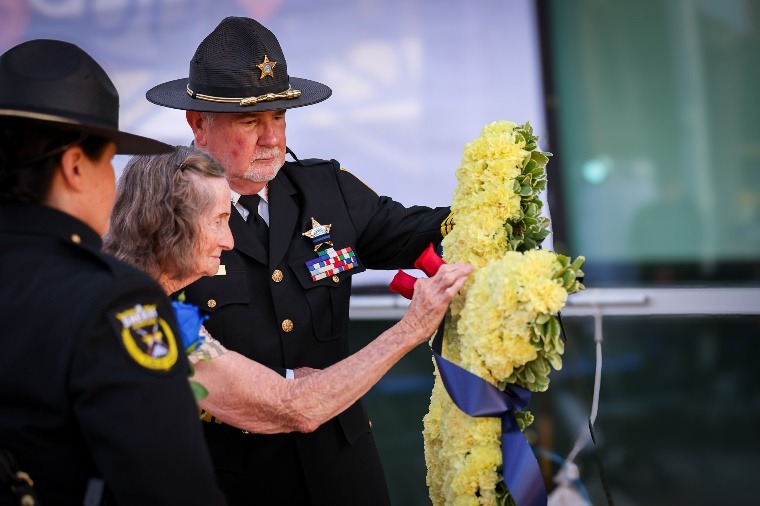 A police officer helps an elderly woman place a wreath of yellow flowers with blue ribbons during a memorial ceremony, while another officer stands nearby