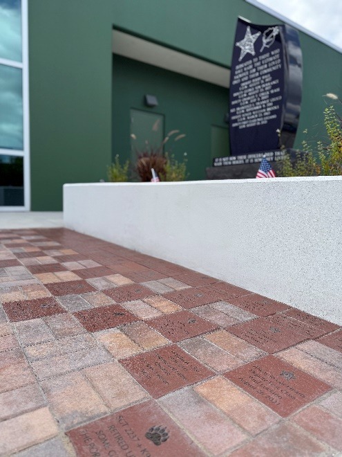 Engraved bricks form a walkway leading to a black stone memorial plaque with small American flags beside it, set against a green building with large windows