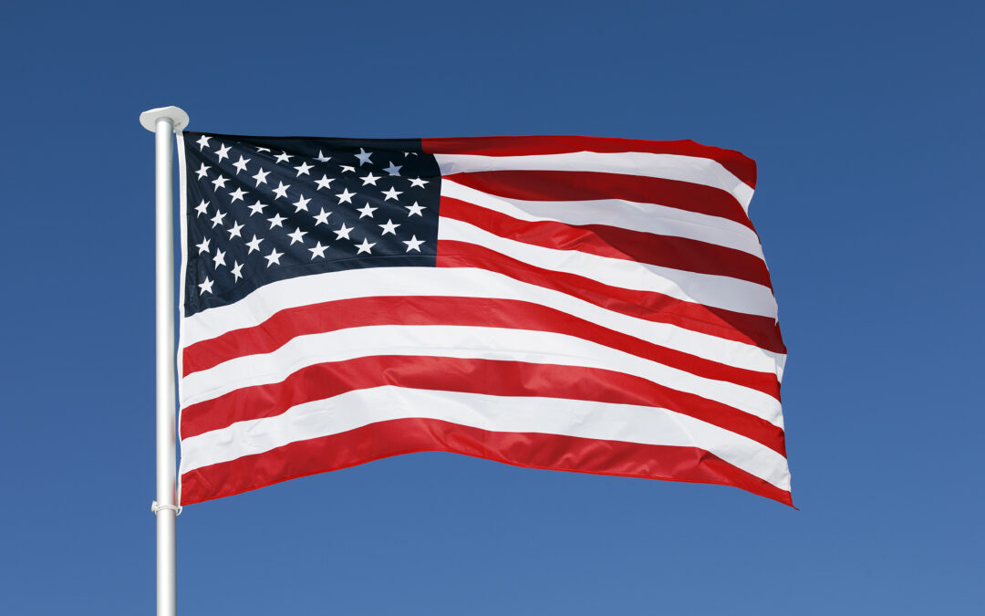 The United States flag with red and white stripes and a blue field of white stars waves in the wind against a clear blue sky