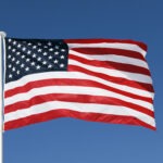 The United States flag with red and white stripes and a blue field of white stars waves in the wind against a clear blue sky