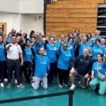 A large group of people in matching blue shirts pose together inside a gymnasium, smiling and raising their index fingers as a gesture of unity and celebration Some people are kneeling in the front row