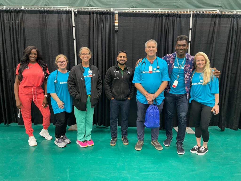 A group of seven people, some in blue AdventHealth shirts and others in scrubs or athletic wear, stand side by side smiling in front of a black curtain on a green floor